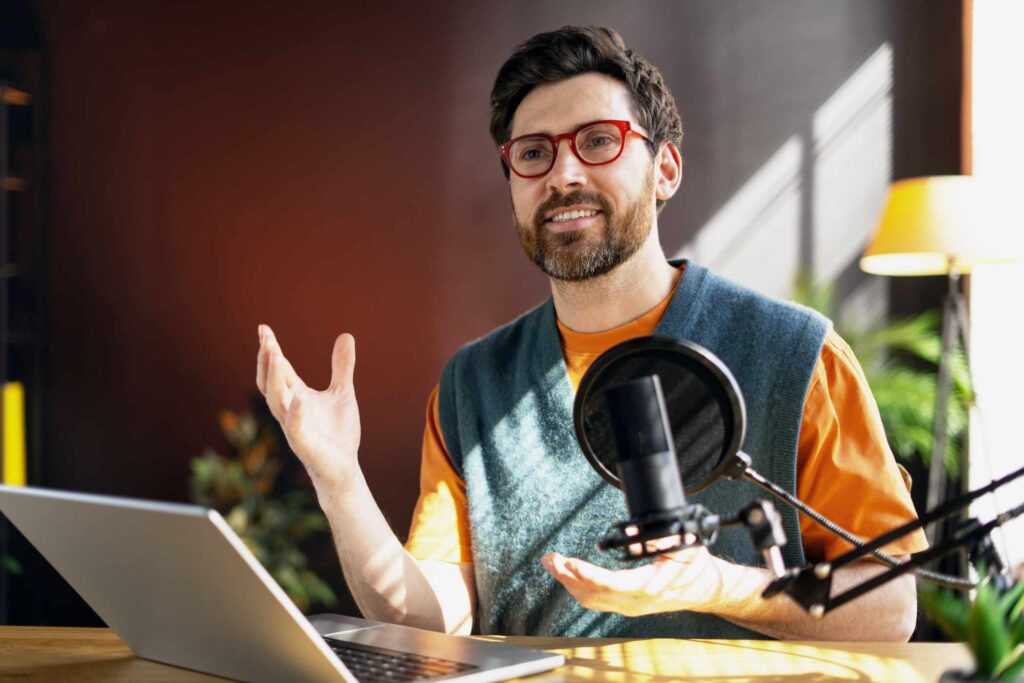 Smiling podcaster gesturing while recording an audio podcast, using a laptop and a professional studio microphone in a home studio setup