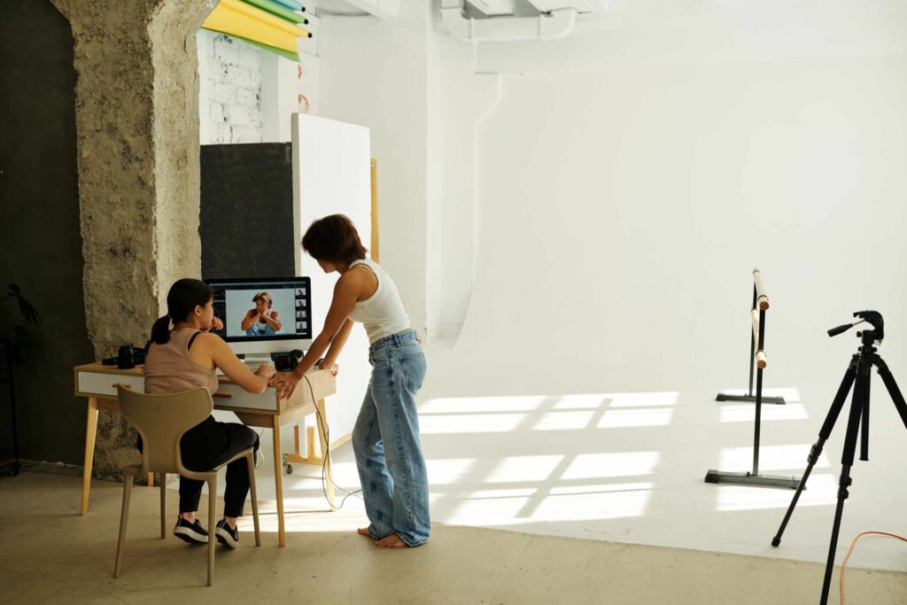 Two young intercultural women in casualwear discussing new shots on computer screen after photo session while one of them sitting on chair