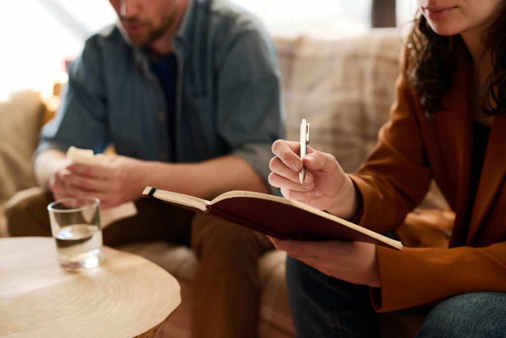 Close-up of young psychologist making notes during conversation with man at session